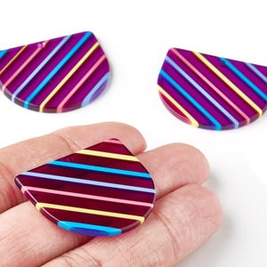 May include: Three purple, semi-circular beads with colourful diagonal stripes in blue, pink, yellow, and white. One bead is held in a hand. These beads are likely for jewellery making or crafting.