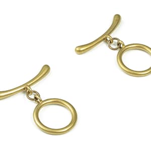 May include: Two gold-toned metal toggles with a ring attached to each. The toggles are shaped like a curved bar with a rounded end. The rings are circular and have a smooth finish.