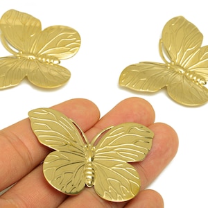 May include: Three gold-colored butterfly charms with detailed wing patterns. One butterfly is held in a hand, while the other two are displayed on a white background. These charms are suitable for jewelry making or craft projects.