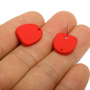 以下が含まれることがあります： 鮮やかな赤色のスタッドピアス。各イヤリングは、上部に小さな穴があり、裏側に銀色のポストが付いた、丸みを帯びた非対称の形状をしています。イヤリングは手の上に表示されています。