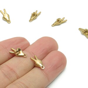 Può includere: Primo piano di diversi piccoli ciondoli dorati a forma di mani che fanno il segno della pace. I ciondoli hanno un anello per attaccarli a gioielli o altri oggetti artigianali. Alcuni ciondoli sono sulla mano di una persona.