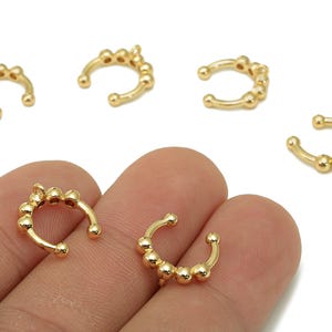 Puede incluir: Puños de oreja dorados con un diseño de cuentas. Los puños de oreja tienen forma curva, con pequeñas cuentas redondas a lo largo de la curva. Varios puños son visibles, algunos descansando sobre una mano.