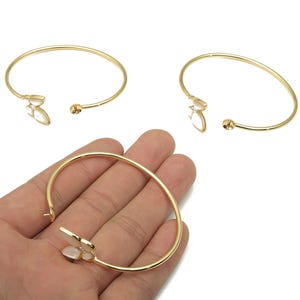 May include: Gold-colored open cuff bracelets with a butterfly design. The bracelets have a delicate, thin band and a small, round ball on one end. One bracelet is being held in a hand.