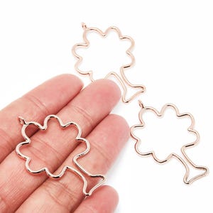 May include: Three rose gold tree-shaped charms. Each charm features a tree outline with a scalloped crown and a slender trunk. The charms are displayed against a white background.