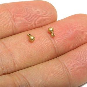 Può includere: Due piccoli elementi di gioielleria dorati, ciascuno con una forma a perla arrotondata e un anello per il fissaggio ad altri componenti. Gli elementi sono mostrati su uno sfondo color carne, evidenziando le loro dimensioni e i dettagli. Questi sono probabilmente utilizzati nella creazione di gioielli.