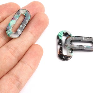 Peut inclure: Deux perles ovales en acrylique avec un motif marbré vert, noir et blanc. Chaque perle a deux trous pour l'enfilage. Une perle est tenue dans une main, tandis que l'autre est posée sur une surface blanche.