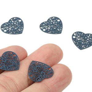 Peut inclure: Gros plan de plusieurs breloques en métal en forme de cœur bleu foncé avec des motifs ajourés complexes et tourbillonnants. Certaines breloques sont éparpillées, tandis que d'autres sont tenues dans une main. Ces breloques décoratives sont idéales pour la fabrication de bijoux ou les projets d'artisanat.