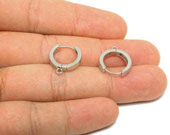 May include: Two silver-colored hoop earrings with small, clear stones set into the outer surface. Each earring has a small loop at the bottom for attaching charms or other decorative elements. The earrings are shown against a neutral background.