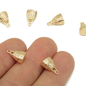 May include: Several small, gold-colored jewelry findings are displayed against a white background. The findings have a bell-like shape with a loop at the bottom and a textured surface. Some are held in a hand.