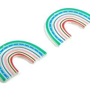 May include: Two colorful rainbow-shaped acrylic charms with a green, blue, pink, red, yellow, and black striped design. The charms have white polka dots on the outer edge.
