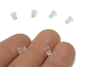 May include: A close-up of several clear, small earring backs. The earring backs are made of a translucent material and are cylindrical with a flat base. Some are held between fingers.