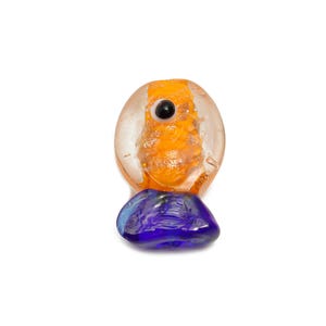 May include: A unique glass art piece featuring a clear, oval-shaped top with orange and gold accents and a black dot. It rests on a rounded, deep blue base. The artwork is set against a white background.