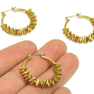 May include: Gold-colored hoop earrings with a unique design. The earrings feature a series of small, rectangular accents around the hoop. One earring is held in a hand, with two others visible in the background. The background is white.