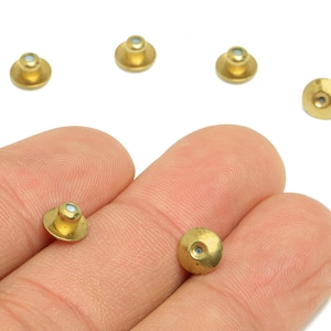 May include: Several small, gold-colored metal studs with a flat, circular base and a small, light-colored center. The studs are scattered on a white background, with some held in a person's hand.