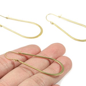 Könnte beinhalten: Goldfarbene, tropfenförmige Ohrringe. Die Ohrringe haben ein schlichtes, längliches Design mit Hakenverschluss. Das Bild zeigt zwei Paar Ohrringe, ein Paar wird in einer Hand gehalten.