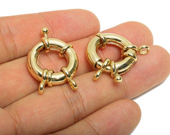 May include: Two gold-colored, round jewelry clasps. Each clasp has a circular body with a hinged opening and a small, decorative ball on the end. The clasps are held in a hand.