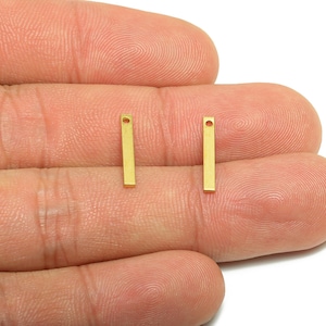 May include: Two small, rectangular gold-colored metal bar charms with a hole at the top. The charms are displayed on a person's hand, against a white background. These charms are suitable for jewelry making.