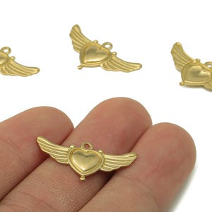 Könnte beinhalten: Goldfarbene herzförmige Anhänger mit angebrachten Flügeln. Die Anhänger haben oben eine kleine Schlaufe zum Befestigen an Schmuck. Das Bild zeigt mehrere Anhänger, von denen einer in der Hand einer Person gehalten wird.