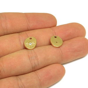 Puede incluir: Dos colgantes de joyería dorados de forma ovalada. Un colgante presenta un diseño de estrella con una piedra central transparente. Ambos colgantes tienen un pequeño agujero en la parte superior para ensartar. Los colgantes se muestran sobre un fondo neutro.
