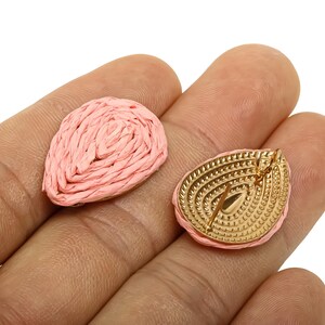 May include: Two pink raffia teardrop-shaped earrings. One earring is face up, showing the woven texture. The other earring is face down, revealing a gold-coloured metal backing with a post for piercing. The earrings are held in a hand.