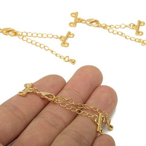 Puede incluir: Extensiones de collar doradas con cierre de barra en T y cadena. Las extensiones tienen un pequeño cierre de mosquetón y una cadena con una pequeña bola en el extremo. Una extensión se sostiene en una mano.
