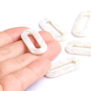 May include: Several white oval-shaped links with a pearlescent finish are scattered on a white surface. One link is held in a hand. These links could be used for jewelry making or other craft projects.