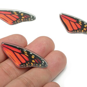 May include: Three monarch butterfly wing-shaped charms. The wings are orange with black and yellow markings, and a clear, glossy finish. One charm is held in a hand. These charms are suitable for jewellery making or craft projects.
