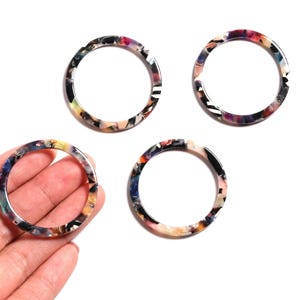 May include: Four colourful acrylic circle charms with a marbled pattern. The charms are in shades of black, white, pink, yellow, and blue. One charm is held in a hand. The charms are on a white background.