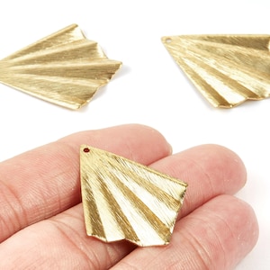 Op de afbeelding: Drie goudkleurige, waaiervormige metalen bedels met een getextureerde afwerking. Eén bedel wordt in een hand gehouden. Elke bedel heeft een klein gaatje om aan sieraden of andere knutselwerkjes te bevestigen. De bedels hebben een geometrisch ontwerp.