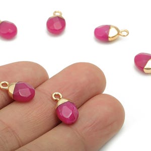 May include: Several faceted, magenta-colored gemstone pendants with gold-colored caps and loops. Some pendants are scattered, while others are held in a hand. The pendants are small, teardrop-shaped, and have a polished finish.