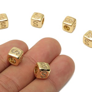 May include: Gold-colored cube beads with the letter "S" on each side. The beads have a hole for stringing and are embellished with small, clear crystals. The beads are shown against a white background.