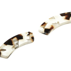 Może przedstawiać: Dwa brązowe i białe spinki do włosów z imitacji żółwia. Spinki są zakrzywione i mają gładkie, błyszczące wykończenie.