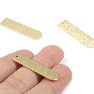 以下が含まれることがあります： テクスチャ加工された表面と丸みを帯びた端を持つ、金色の長方形の金属チャームが3つ。1つのチャームが手で持たれています。各チャームには端に小さな穴があり、ジュエリー作りや工芸品用であることが示唆されています。