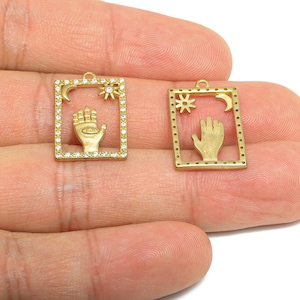 May include: Two gold-colored rectangular pendants. One pendant has a hand with an eye in the palm, a moon, and a sun, framed with small clear stones. The other pendant has a hand, a moon, and a sun.