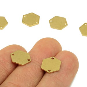 Op de afbeelding: Verschillende kleine, goudkleurige, zeshoekige metalen bedels met twee of drie gaten voor het maken van sieraden. De bedels worden getoond tegen een witte achtergrond, sommige worden in een hand gehouden.
