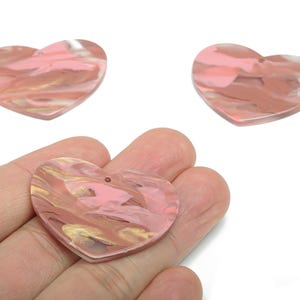 May include: Three heart-shaped acrylic pendants with a marbled design in shades of pink, brown, and gold. One pendant is held in a hand, while the other two are displayed separately. Each pendant has a small hole at the top.