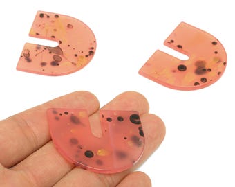 May include: Three translucent, U-shaped, peach-colored gemstone pendants with dark brown and gold inclusions. One pendant is held in a hand. The pendants are likely for jewelry making or crafting.