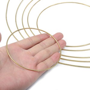 Puede incluir: Una mano sosteniendo un aro de metal dorado. Varios otros aros dorados de diferentes tamaños son visibles en el fondo, lo que sugiere un tema de artesanía o fabricación de joyas. Los aros son delgados y circulares.