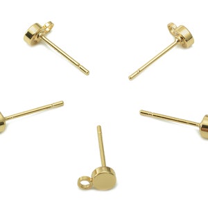 Pode incluir: Uma coleção de brincos de botão dourados. Cada brinco apresenta um pequeno disco redondo com um pino para inserção e uma argola para prender pingentes. Os brincos são exibidos em um fundo branco.