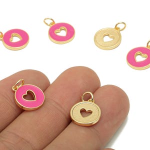 May include: Gold-colored metal charms with a heart-shaped cutout. Some charms have a bright pink enamel border. The charms are round with a small loop at the top for attaching to jewelry. The image shows several charms.