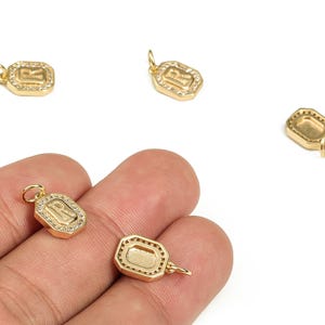 Peut inclure: Charm pendentif doré avec la lettre "R" à l'intérieur d'une forme rectangulaire, entouré de petits cristaux transparents. Les breloques sont présentées en gros plan, certaines étant tenues dans une main.