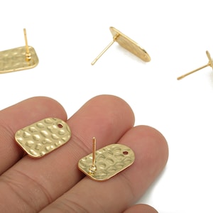 Puede incluir: Pendientes de botón dorados con textura martillada. Los pendientes tienen forma rectangular con bordes redondeados y un poste para la inserción. Se ven varios pendientes, algunos con el poste visible y otros sin él.