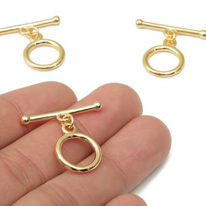 以下が含まれることがあります： 金色のトグルクラスプ。ジュエリー作りに最適です。クラスプはバーとリングで構成され、シンプルでエレガントなデザインです。画像には3つのクラスプがあり、1つは手に持たれています。