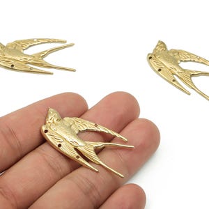 Könnte beinhalten: Drei goldfarbene Metallanhänger in Form von Schwalben. Die Anhänger sind detailliert mit Flügel- und Schwanzfedern und haben kleine Löcher zum Befestigen an Schmuck oder anderen Bastelarbeiten. Die Schwalben sind auf weißem Hintergrund abgebildet.