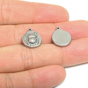 May include: Two small, silver-colored pendants. One pendant features a detailed scarab beetle design within a circular frame. The other pendant is a plain, smooth circle. Both have a small loop at the top for attaching to a chain or other jewelry.