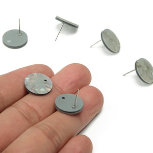 Op de afbeelding: Grijze en iriserende oorstekers met zilveren pinnen. De oorbellen zijn rond met een plat, cirkelvormig ontwerp. Verschillende oorbellen zijn verspreid op een witte achtergrond, sommige worden in een hand gehouden.