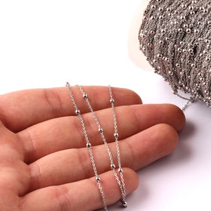 May include: Silver-coloured beaded chain on a white background. The chain is draped across a hand, with a spool of the same chain visible. The chain features small, evenly spaced beads.
