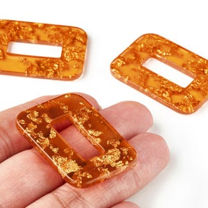 May include: Three rectangular orange resin beads with gold leaf inclusions. Each bead features a rectangular aperture in the centre. The beads are displayed against a white backdrop.