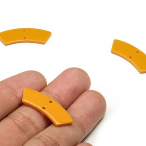 Pode incluir: Três botões curvos amarelo mostarda, cada um com dois furos. Um botão é segurado na mão, enquanto dois outros estão em um fundo branco. Estes botões são para projetos de costura ou artesanato.