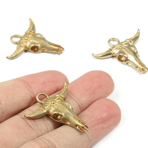 May include: Gold-coloured metal bull skull charms. The charms have a loop for attaching to jewellery. The skulls have detailed horns and facial features. The charms are shown on a white background.